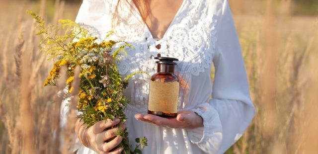 natural-remedies-beautiful-woman-in-a-field A woman holding a bouquet of freshly gathered wild herbs and an amber glass apothecary bottle in a sunny field representing the natural benefits of Chinese herbal medicine