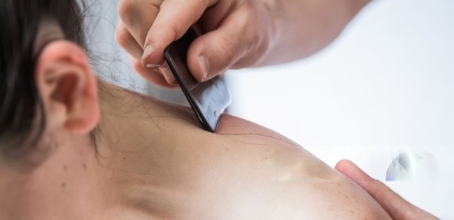 Practitioner applying targeted press-stroke friction with a smooth scraping tool along the cervical spine and trapezius muscle zone